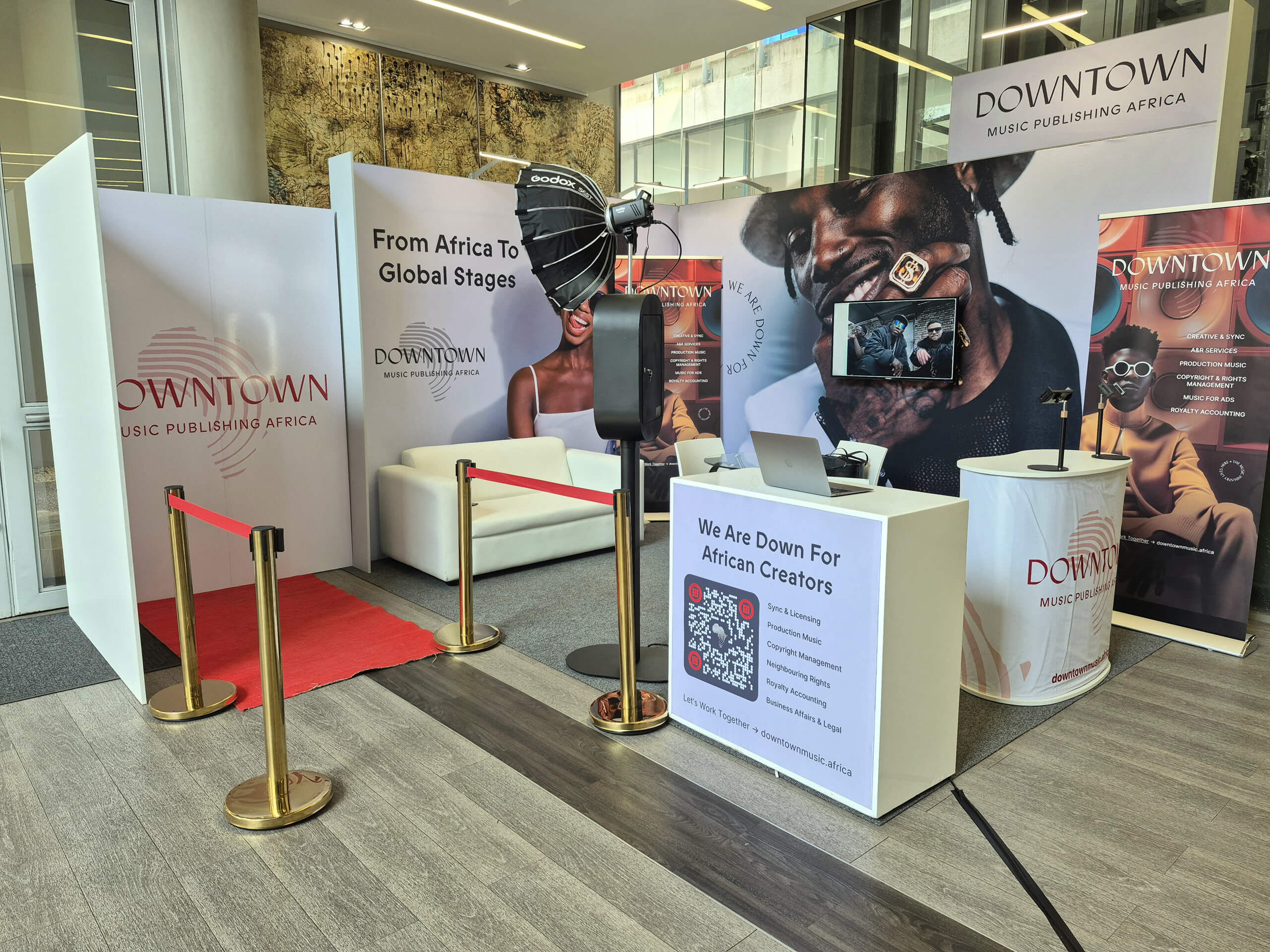 A modern exhibition stand for Downtown Music Publishing Africa features banners, a red carpet, a white sofa, a microphone, a TV displaying musicians, a laptop, promotional posters, and a QR code, all inside a sleek, glass-walled space.