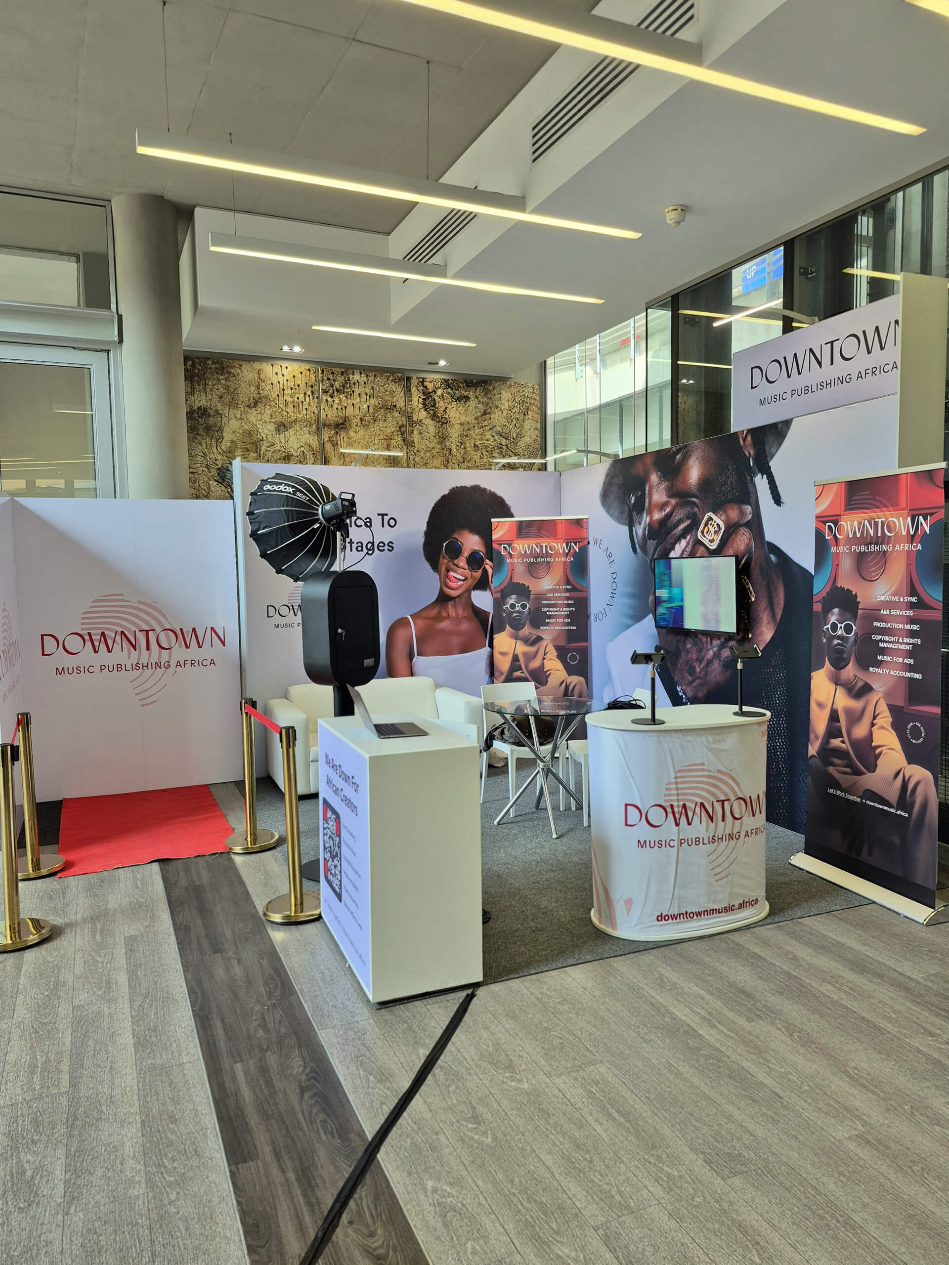 A modern exhibition stand for Downtown Music Publishing Africa features banners with smiling Black musicians, a ring light, a TV screen, a white desk with brochures, and a red carpet. The stand is set in a bright, contemporary room with wood and carpet flooring.