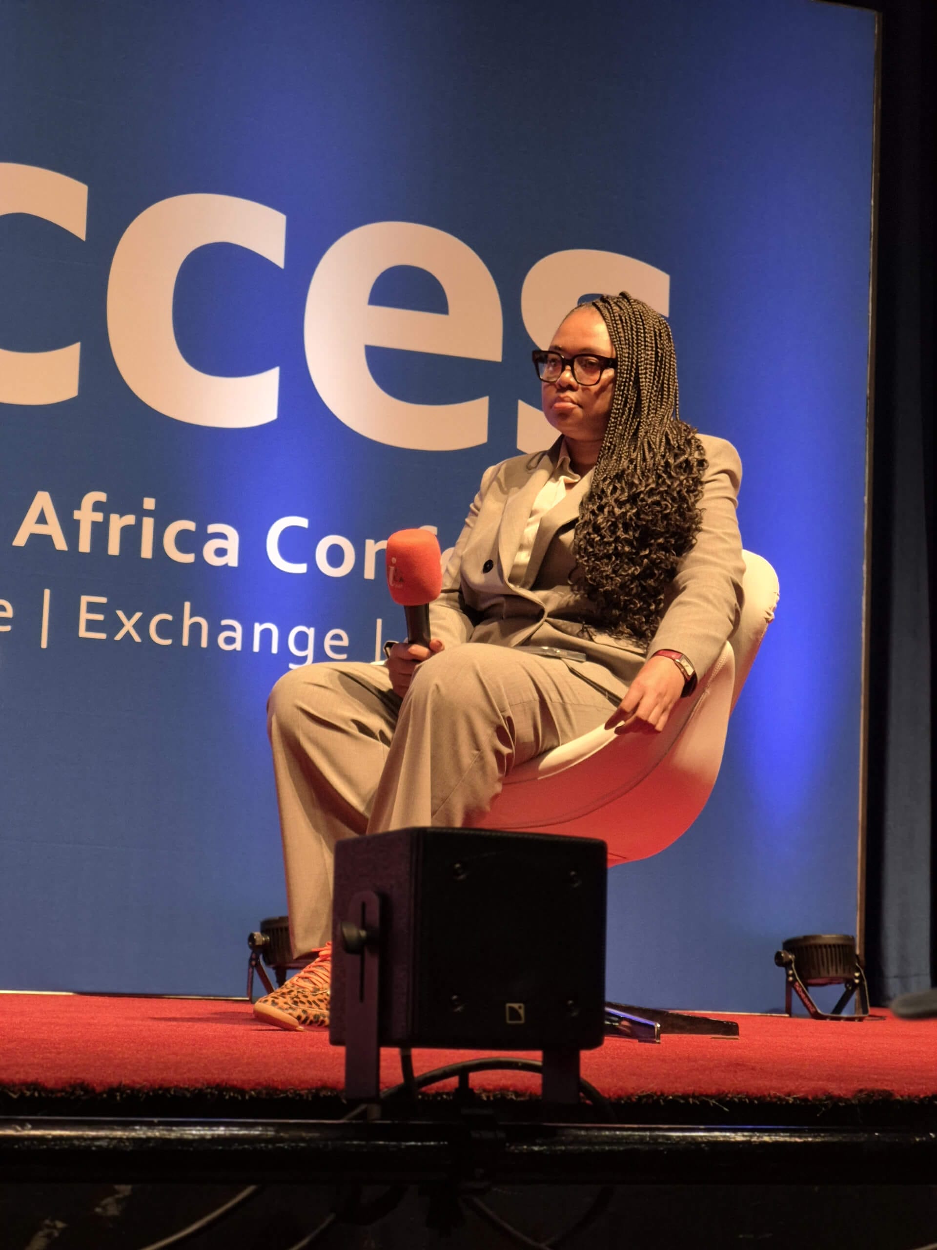 A person in a light grey suit and glasses sits on a white chair holding a microphone. They have long plaits and orange shoes. Behind them is a blue backdrop with partial white text reading “Africa Con… Interact | Exchange |…”.