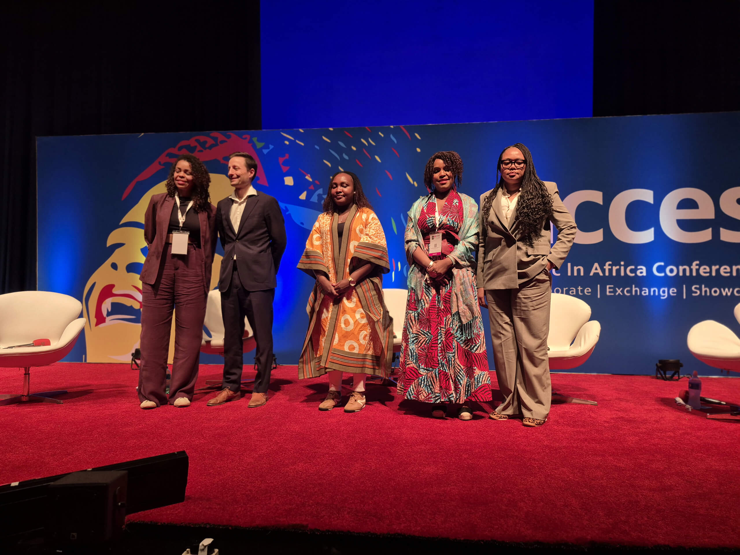 Five people stand on a red-carpeted stage in front of a blue backdrop featuring artwork and the word 