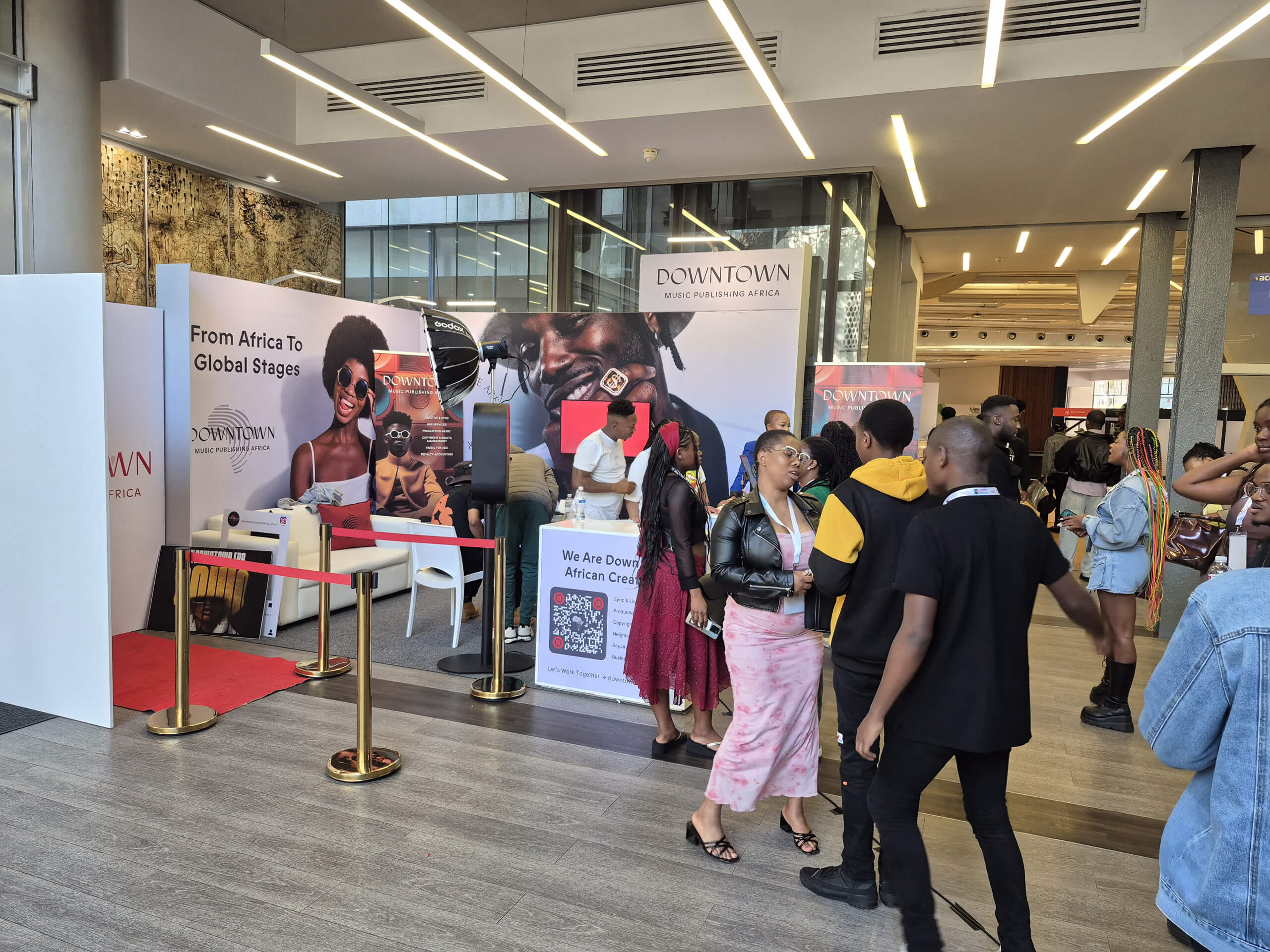A group of people gather at an indoor event near a stand with "Downtown Music Holdings Africa" banners. Some people chat, while others look at displays. The stand features posters, brochures, and ropes, with a modern, well-lit background.
