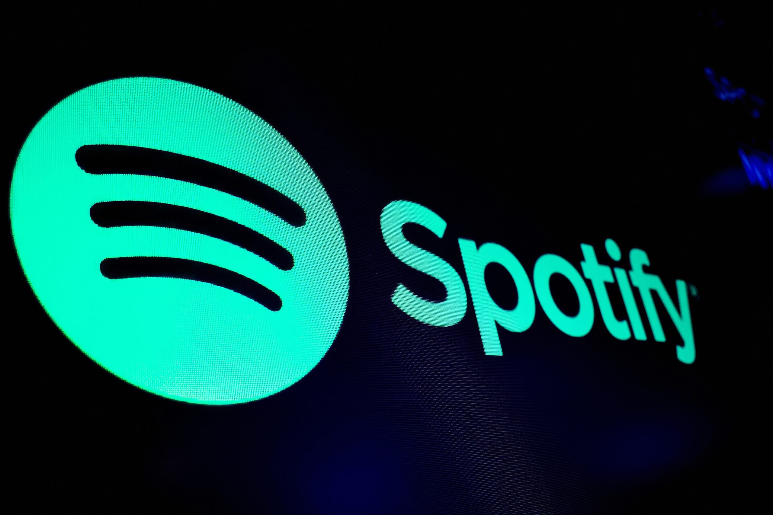 The image shows the Spotify logo: a bright green circle with three curved black lines inside, alongside the word “Spotify” in bold, light green letters, all set against a dark black background. The logo is glowing, creating a vibrant, modern look.