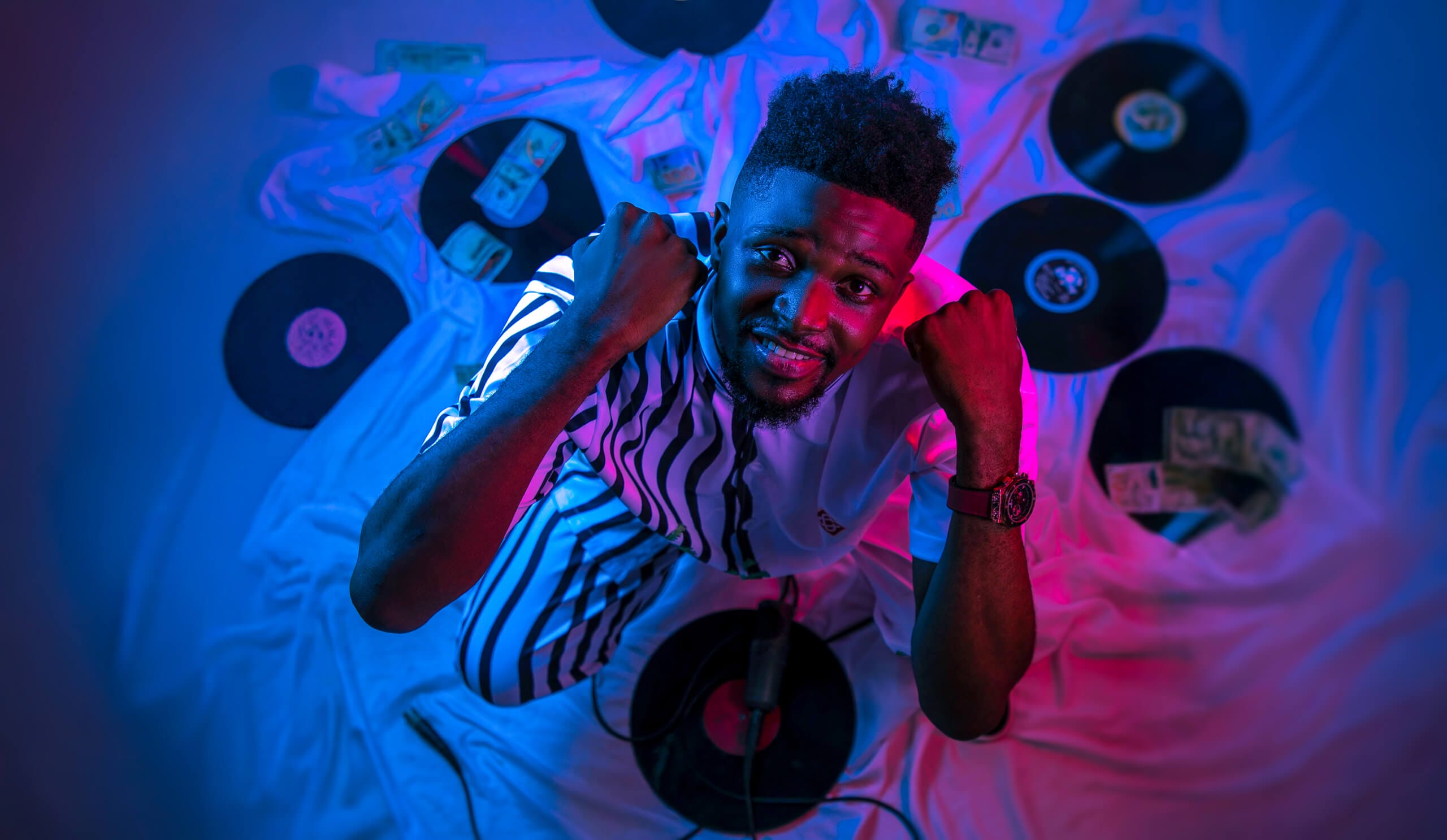 A man in striped trousers and a white shirt kneels on a white sheet, surrounded by vinyl records and scattered cash. He looks up and raises his fists, smiling. The scene is lit with vibrant blue and pink lighting, creating a dramatic, creative atmosphere.
