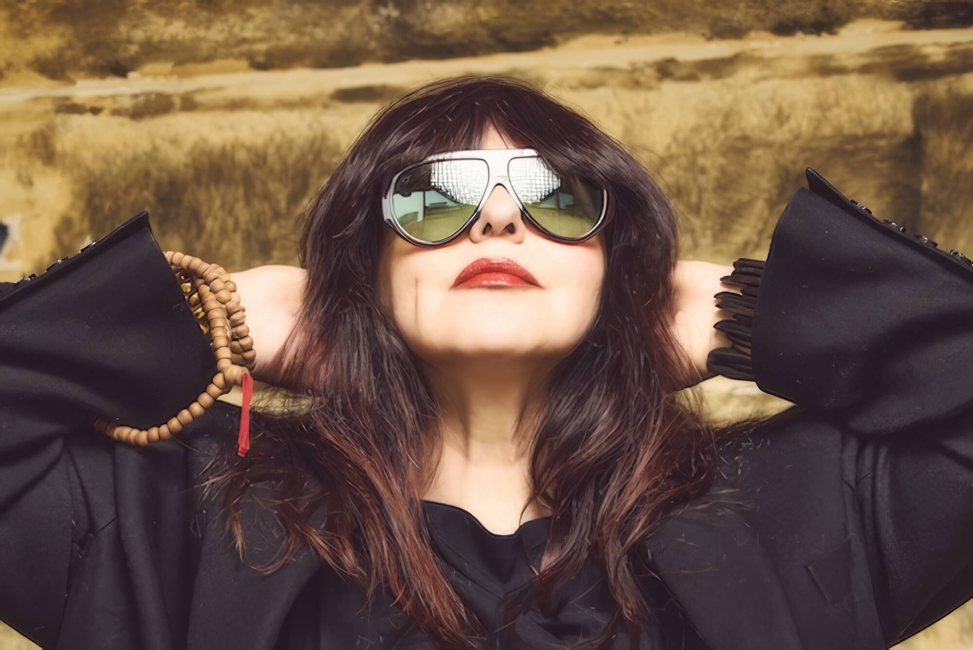 A woman with long, dark hair and red lipstick wears large aviator sunglasses and a loose black outfit. With an introspective depth reminiscent of "Maybe I Already Know" by Hanna Haïs, she poses confidently before a blurred urban brown wall.