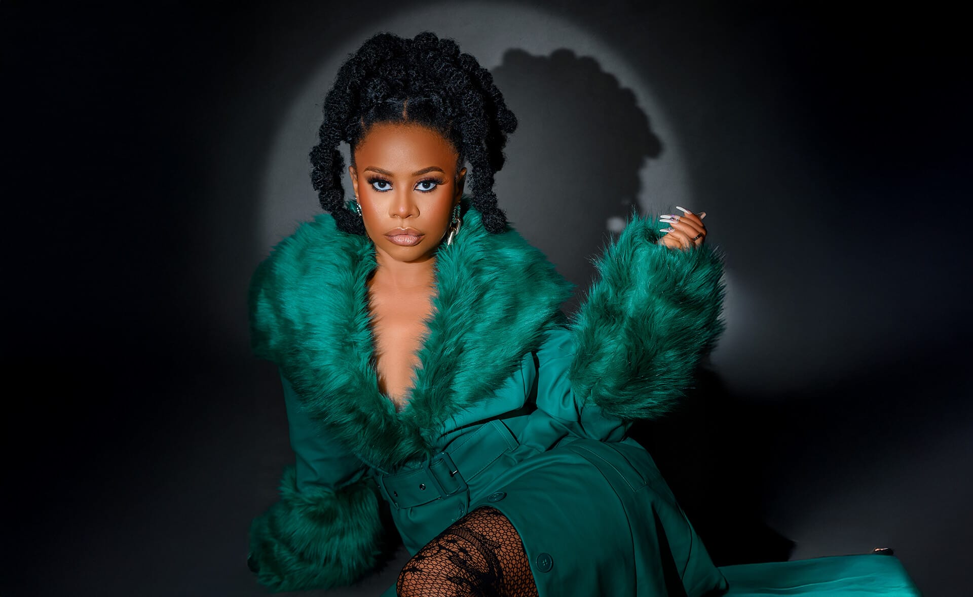 A woman, channelling Energy Goddess vibes, sits against a dark background in a deep green coat with a faux fur collar. With styled natural hair and bold blue eye make-up, she brings Afro-Dancehall flair to the scene beneath dramatic lighting.