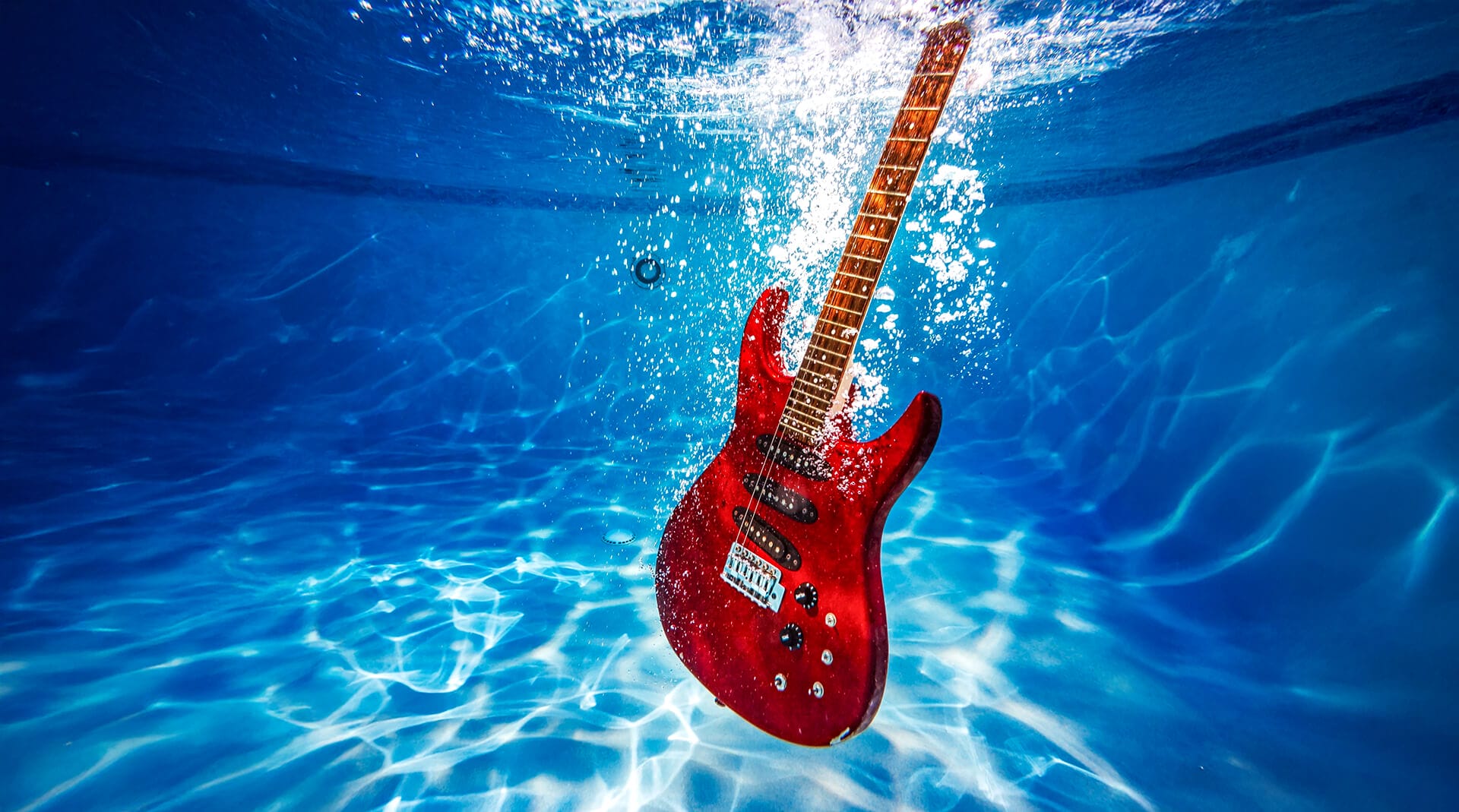 A chilled guitar, submerged underwater in a swimming pool, is surrounded by bubbles and ripples. Sunlight filters through, casting wavy patterns on the blue pool floor as the strings and knobs remain clearly visible.