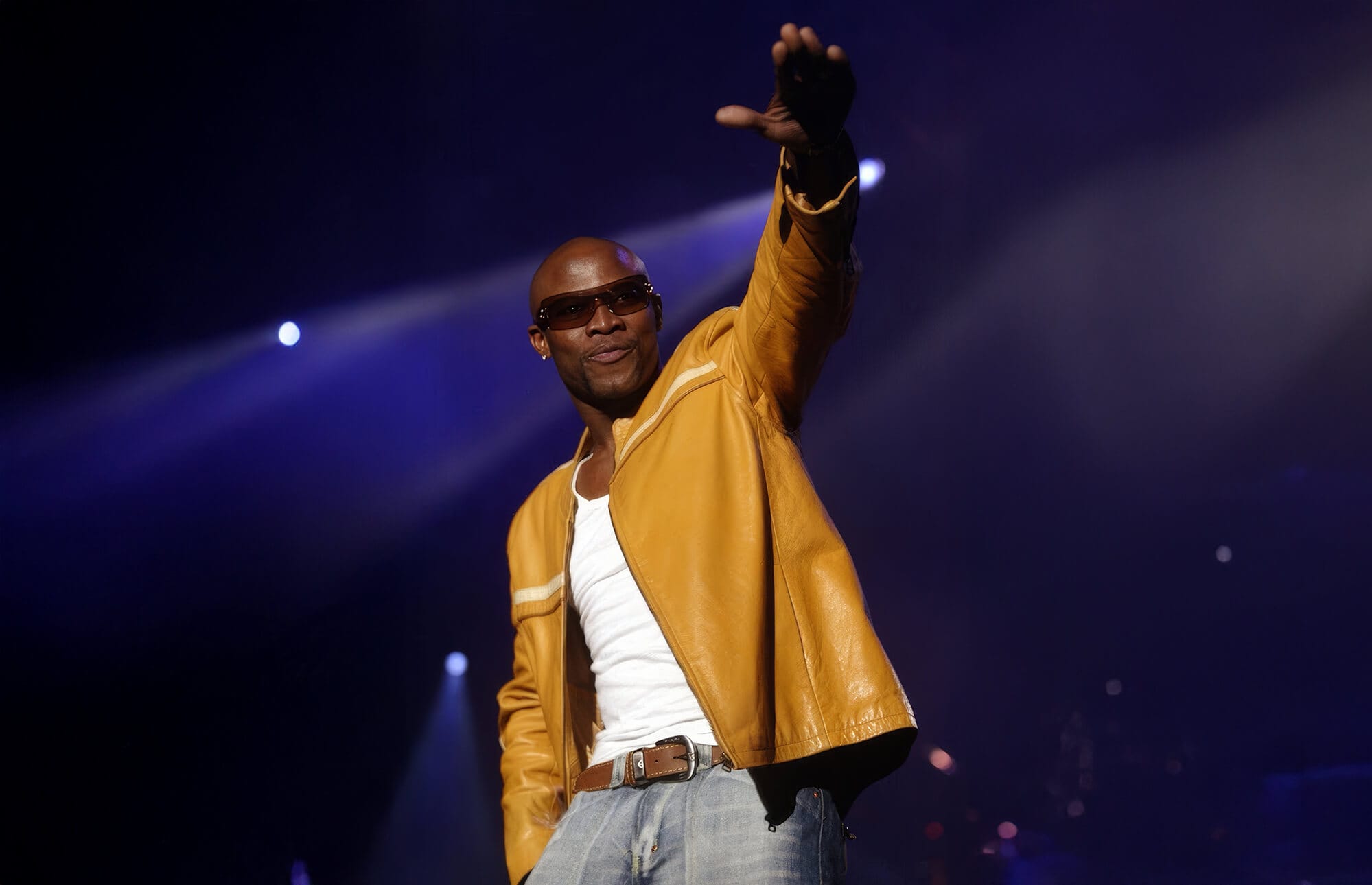 A man wearing sunglasses, a yellow leather jacket, white shirt, and jeans stands on stage under bright spotlights. Smiling confidently, he raises his right arm forward in true Nkalakatha style—a nod to South African music and its rite of passage for many artists.