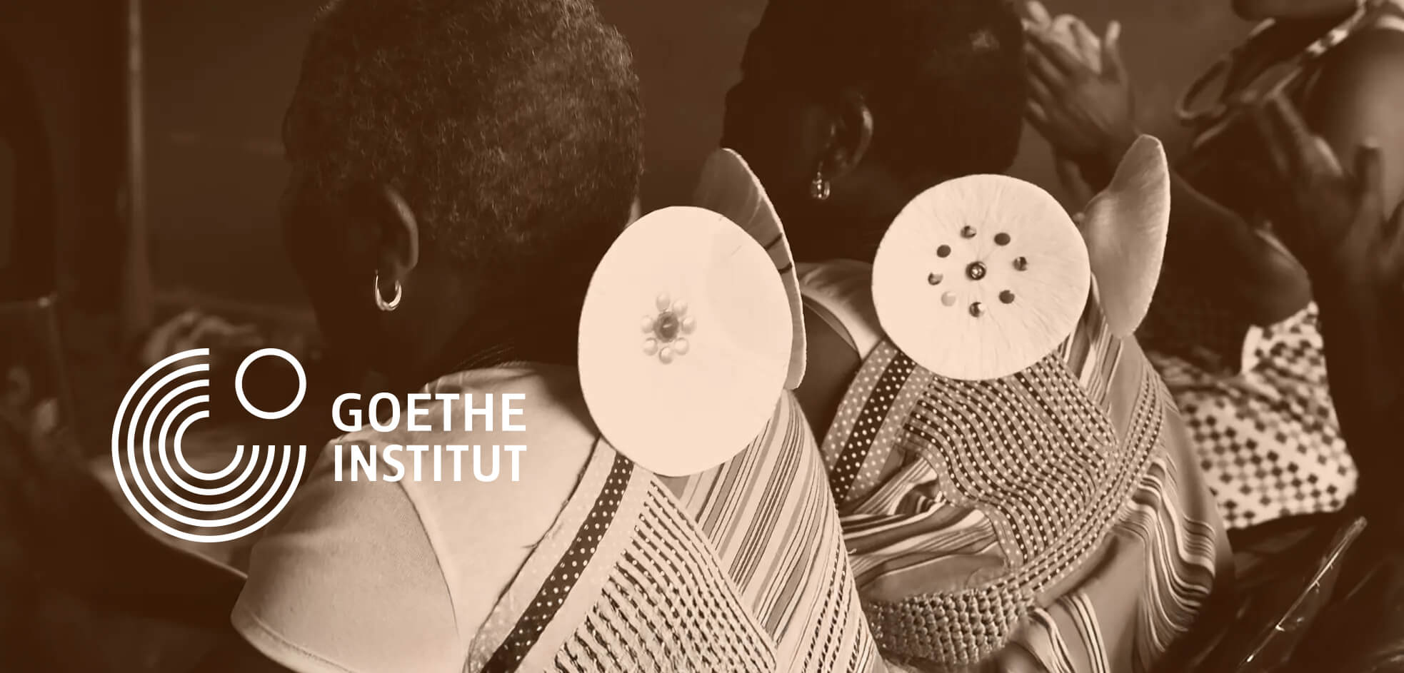 Two women seen from behind wear traditional, patterned clothing with circular white adornments on their backs. A sepia tone covers the image. On the left, the Goethe-Institut logo and text are visible. Other people’s hands are blurred in the background.