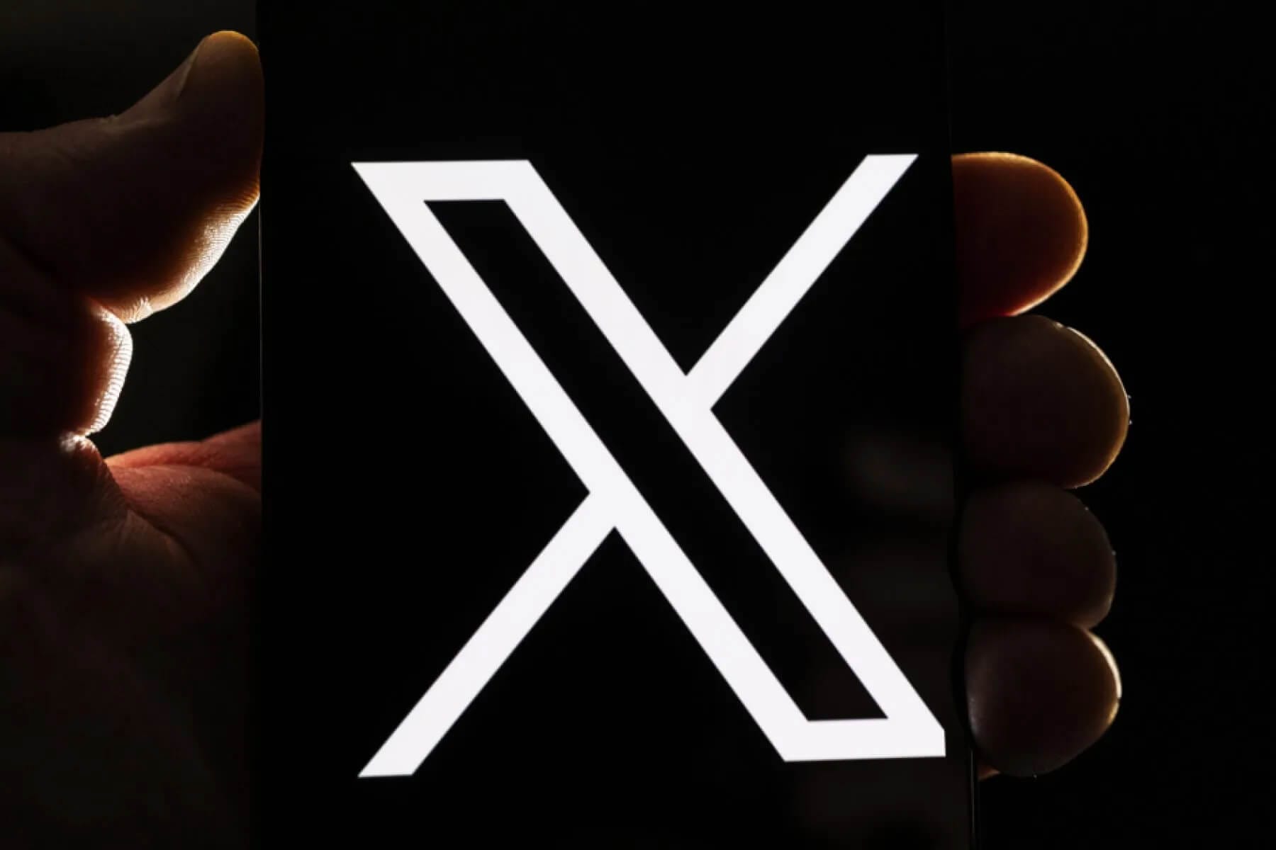 A hand holds a smartphone displaying a bold, white, stylised “X” on a black screen. The background is dark, highlighting the illuminated letter and the silhouette of the hand and fingers around the phone.