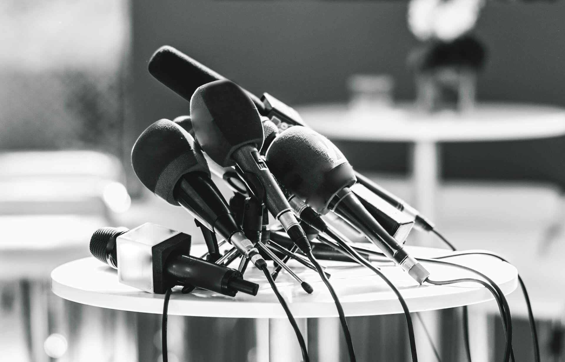 A cluster of microphones and audio recorders, some with foam covers, are arranged on a round white table. The black-and-white image hints at a press conference, possibly following a European Commission approval in a city centre x Virgin setting.