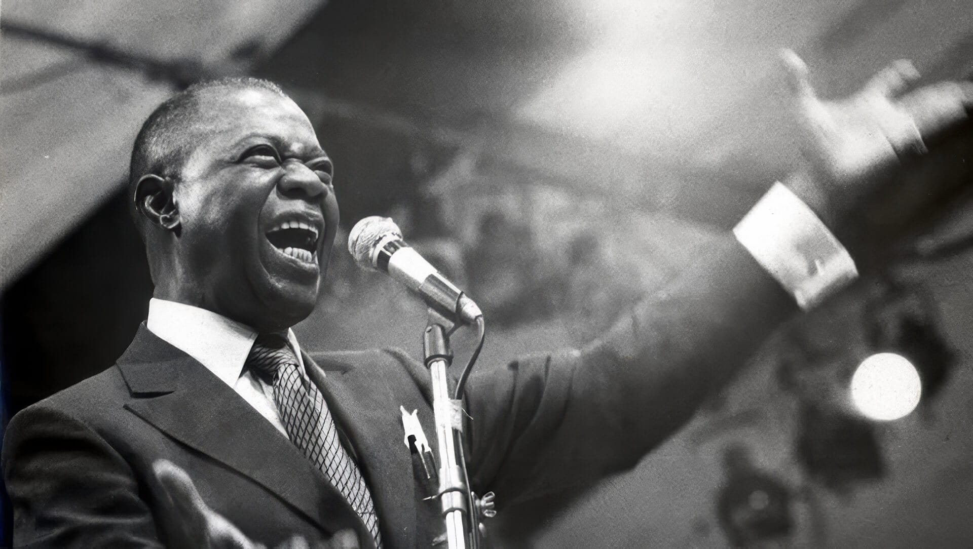 A Black man in a suit passionately sings into a microphone on stage, arms spread wide. He is smiling broadly, eyes closed, conveying joy and emotion. The background is out of focus with bright stage lighting highlighting his face.