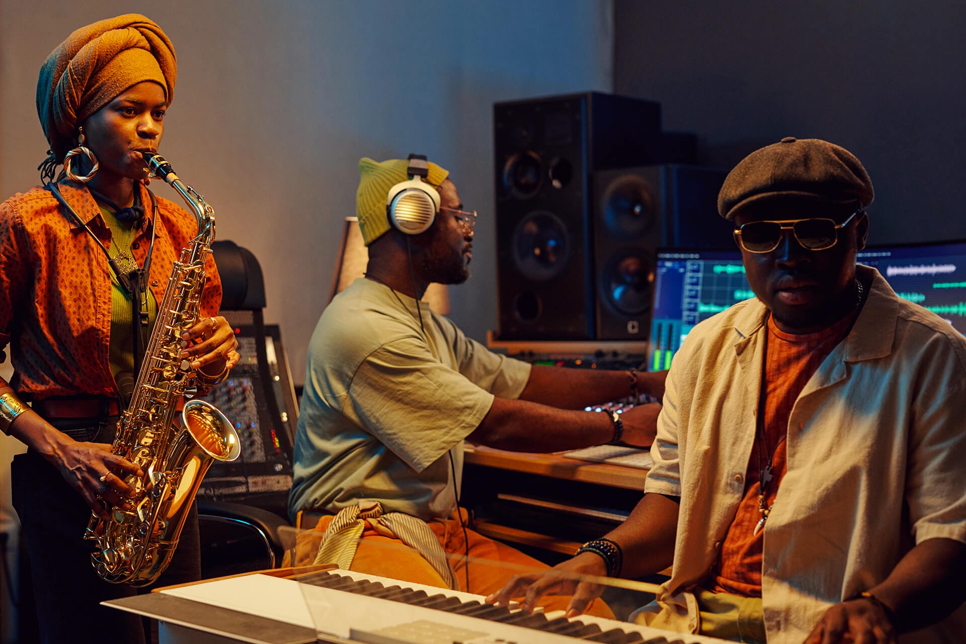 Three Black musicians in a recording studio: a woman plays saxophone, a man in headphones works at a mixing desk, and another man in sunglasses and a cap plays keyboard. The room is dimly lit with monitors and audio equipment in the background.