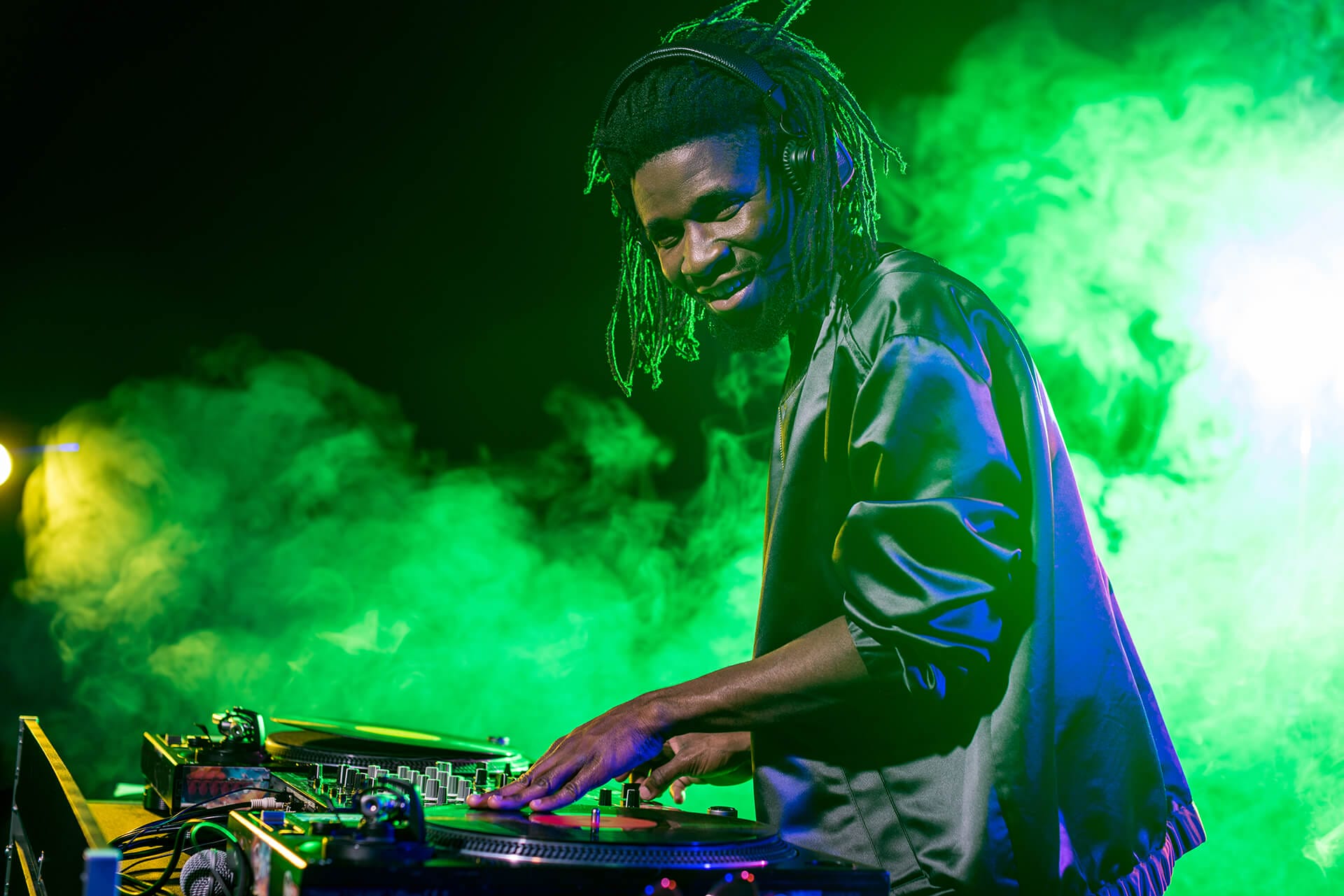 A DJ with long dreadlocks and headphones smiles whilst mixing music on turntables. He wears a shiny black jacket and, representing Nigeria, energises the crowd amid vibrant green stage lights and swirling smoke.