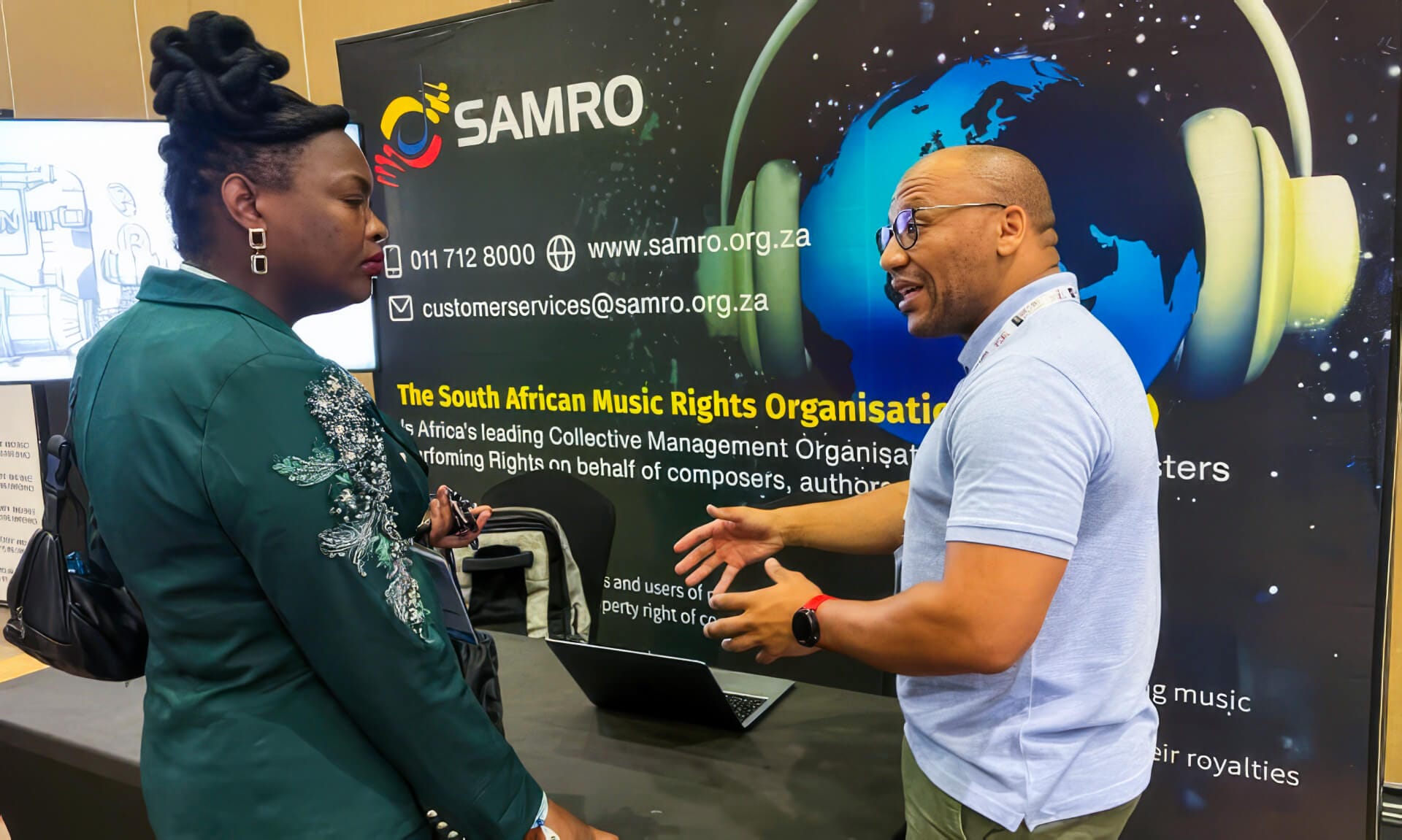 A man and a woman converse in front of a SAMRO stand. The banner highlights “The South African Music Rights Organisation,” music rights, and contact info as they discuss recent SAMRO distribution changes and strategies for better cashflow planning.