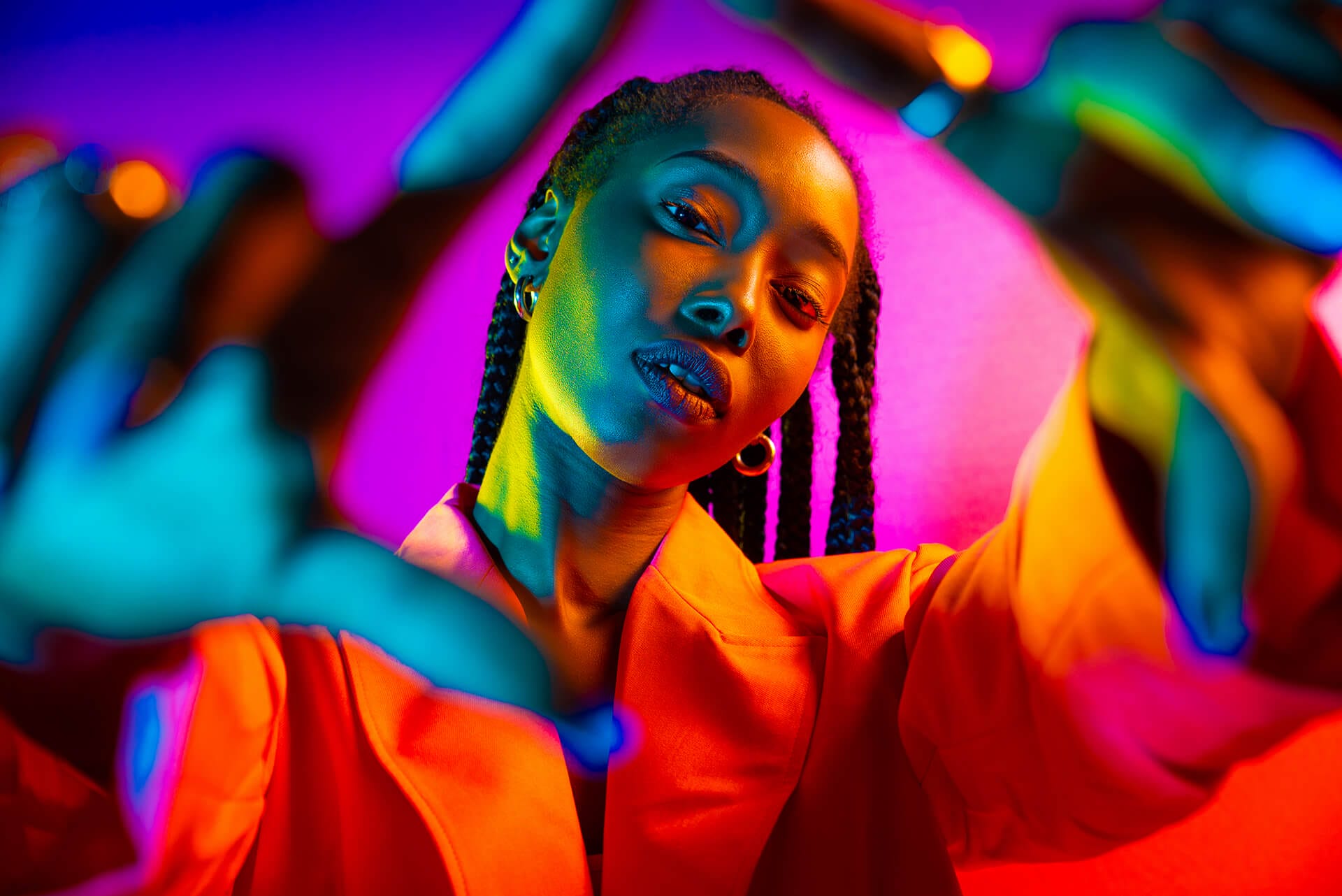 A woman with plaits wears a bright orange blazer and hoop earrings. She poses confidently with her hands framing her face. Vivid blue, pink, and orange lights create a dramatic effect inspired by Africa’s vibrant style against a gradient background.