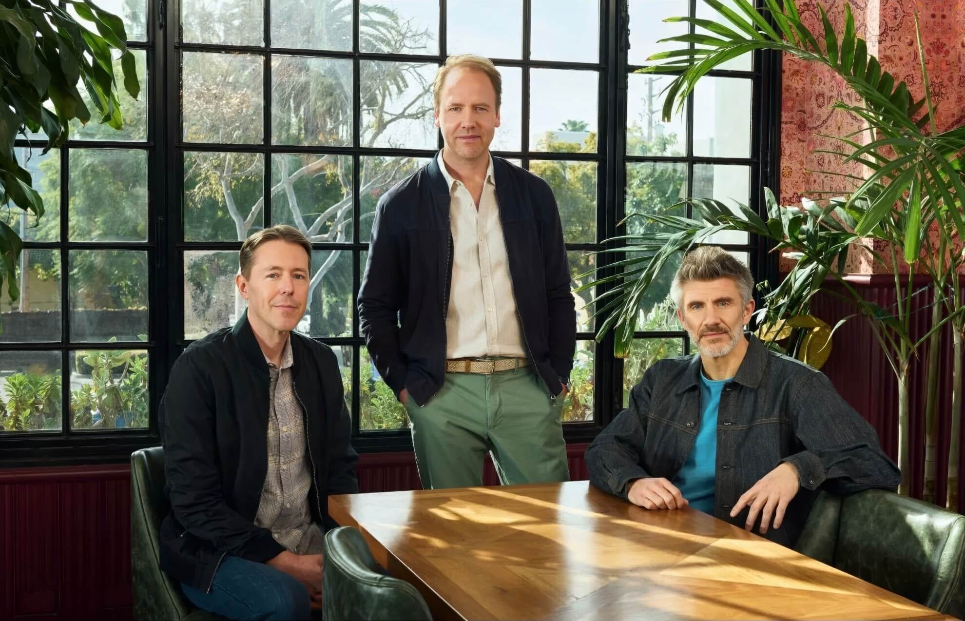 Three men pose indoors by a large sunlit window with palm trees visible outside. One man stands leaning on a table whilst the other two sit on either side. Potted plants fill the room, and all are dressed in casual jackets and shirts, looking at the camera.