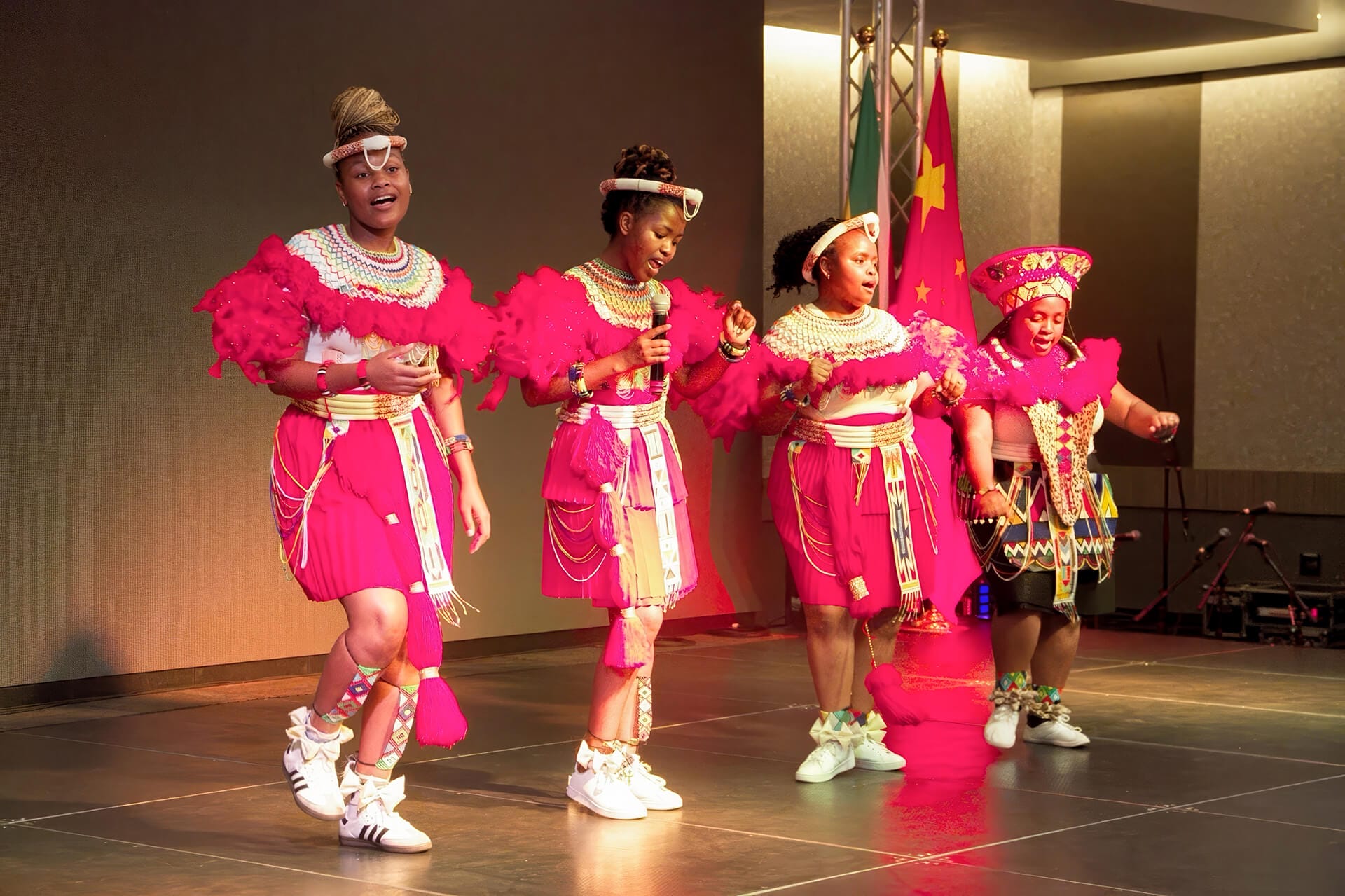 Four women in vibrant pink traditional attire with elaborate beadwork perform a cultural dance on stage, representing eThekwini Arts. They wear white trainers and unique headpieces, with a Cameroon flag in the background under warm stage lighting.