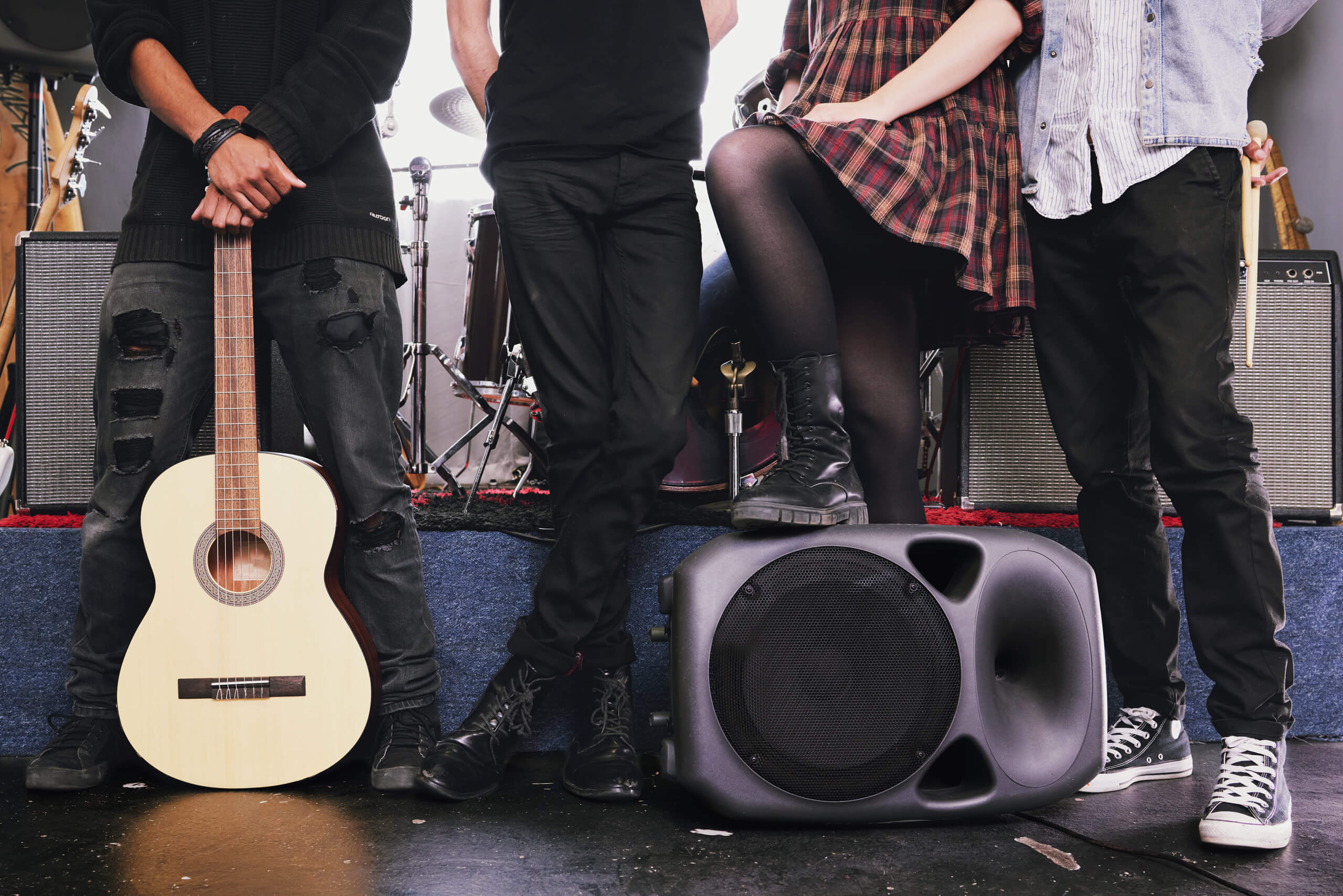 Four people stand side by side in front of music equipment; only their lower bodies are visible. One holds an acoustic guitar, another stands with arms folded, a third has a boot on a speaker, and the fourth wears trainers and rests an elbow on a knee.