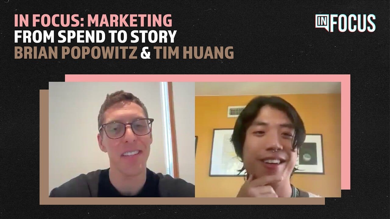 Split-screen video call with two men. Left: Man with glasses, short brown hair, light skin, smiling, white wall in background. Right: Man with long dark hair, tan skin, hand on chin, yellow wall and artwork behind. Title: "IN FOCUS: From Marketing Spend to Story.
