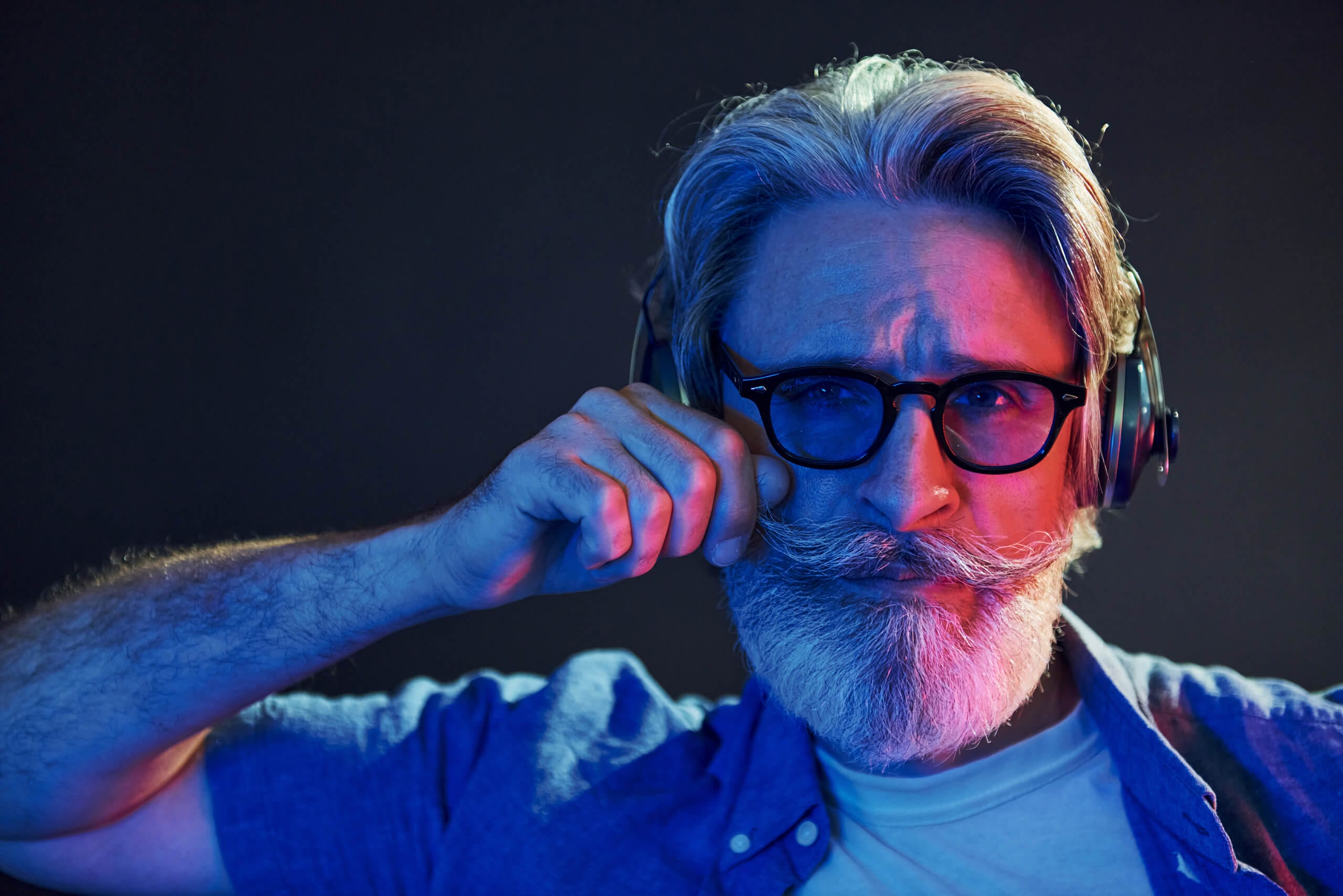 A middle-aged man with grey hair, a thick moustache, and a beard wears glasses and large headphones. He adjusts his moustache with one hand and looks intently at the camera. Blue and pink lighting cast colourful shadows on his face and shirt.