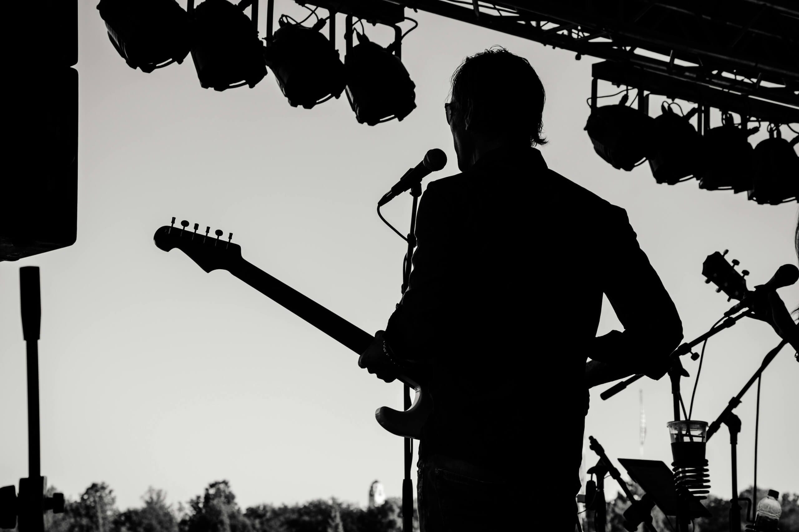A black-and-white photo shows a musician silhouetted on stage, playing an electric guitar and singing into a microphone. Stage lights hang above. Trees and a clear sky are visible in the background, creating a contrast between artist and surroundings.