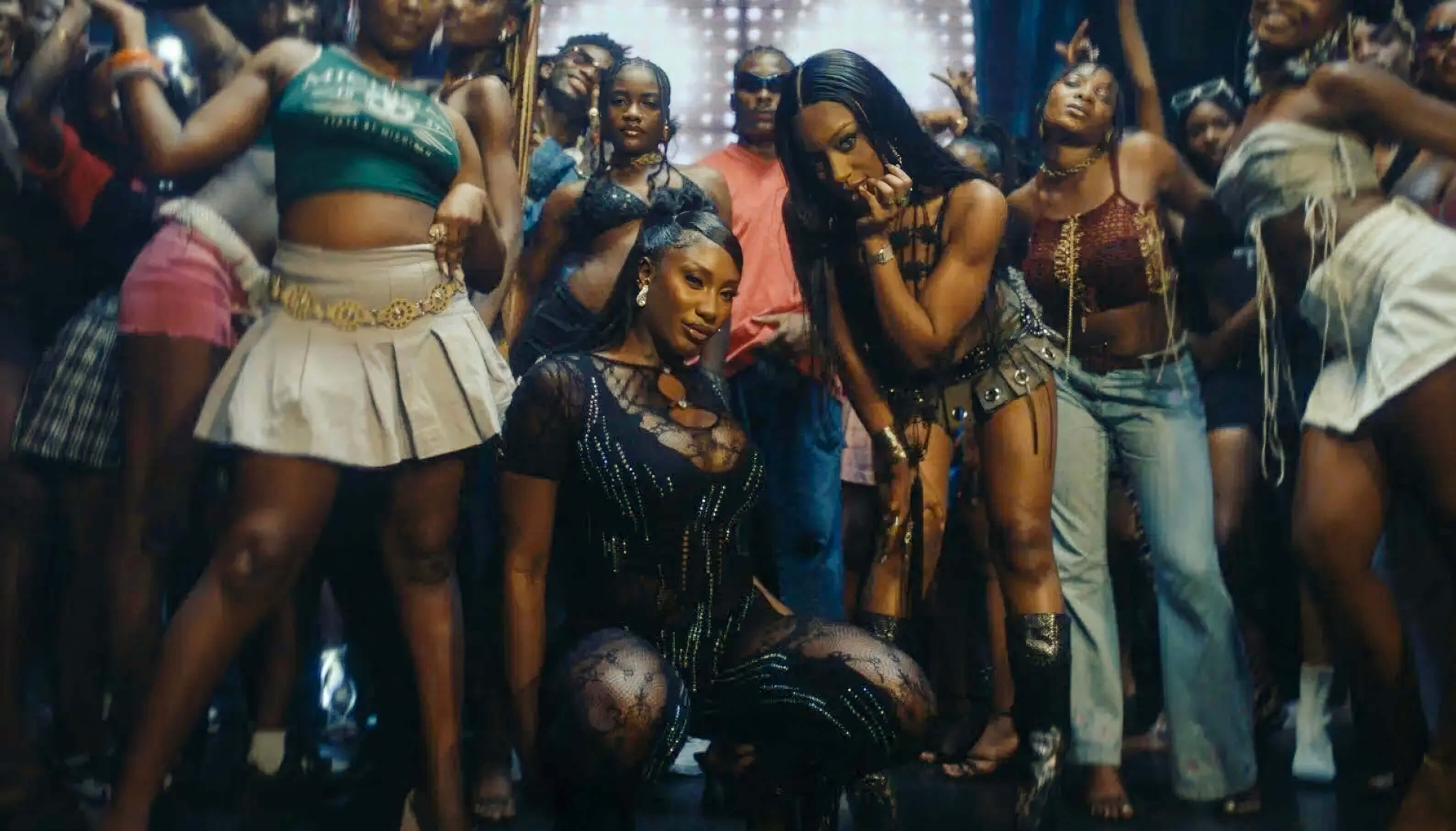 A lively group of young Black women pose and dance at a crowded party, celebrating Africa–diaspora music collaborations. Two women—one kneeling in black mesh, one in a gold chain skirt—lead as others smile and dance under festive lights.