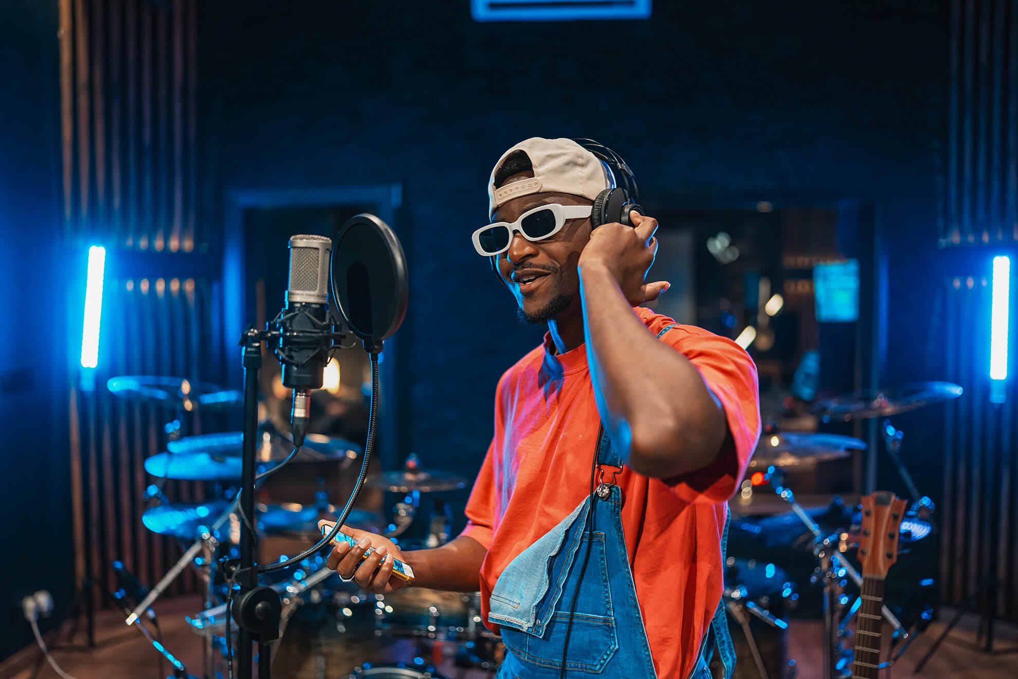 A man wearing white sunglasses, a backwards cap, red shirt, and denim dungarees sings into a studio microphone, holding headphones to his ear. He stands in a music studio with drums, guitars, and blue neon lights—pursuing sustainable music careers.