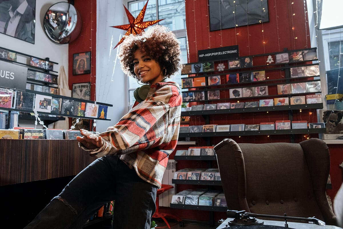 A smiling person with curly hair and headphones around their neck stands in a cosy record shop. Surrounded by hit songs on CD shelves, fairy lights, and a star decor on a red wall, they embody the warm, inviting vibe behind the scenes of music culture.