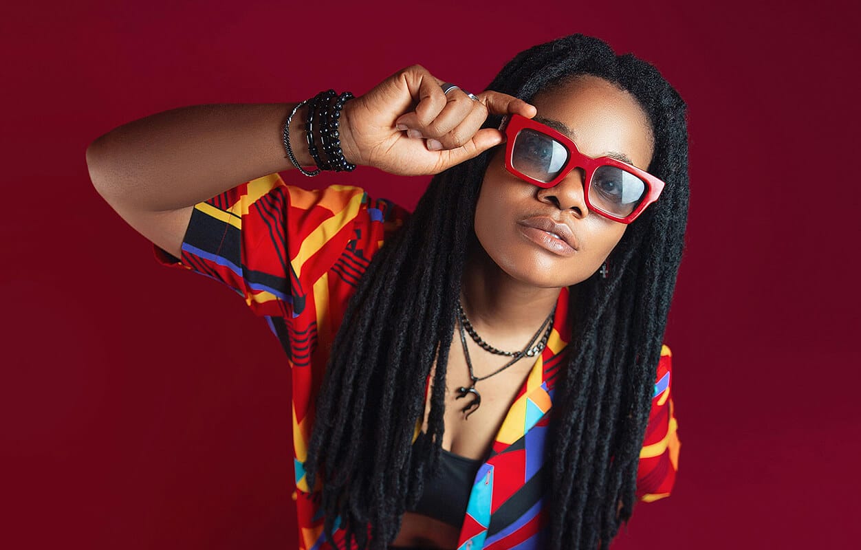A person with long black dreadlocks and dark skin wears red sunglasses, a colourful patterned shirt, black top, and layered necklaces. Posing confidently against a deep red background, they embody the vibrant spirit of the African music industry.